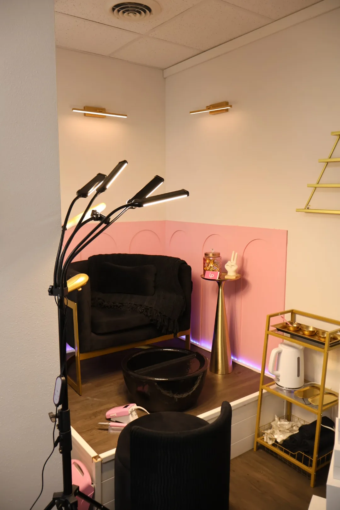 Pedicure corner with a pink wall, a black armchair, a gold side table and a cart with a kettle.