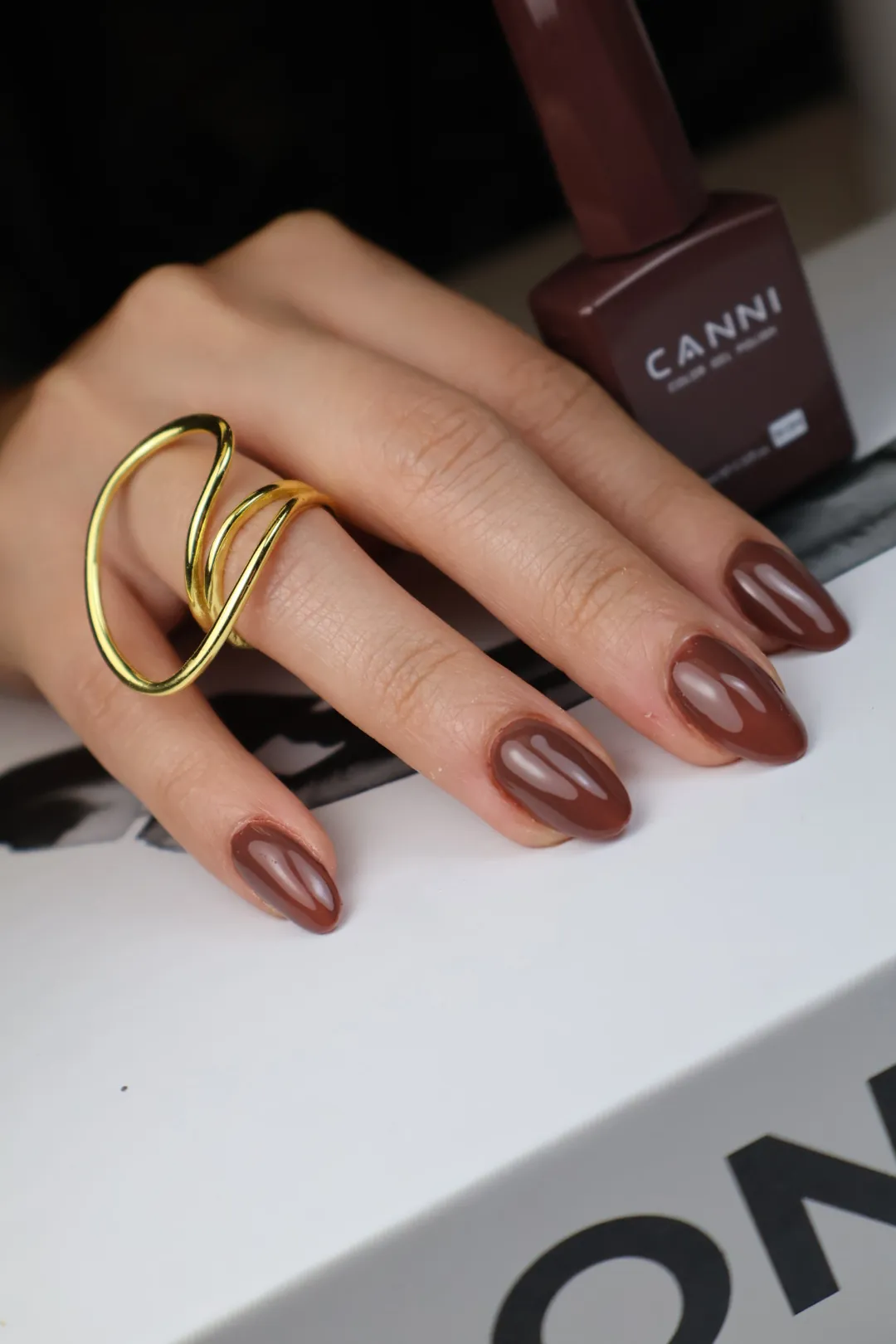 A hand with glossy chocolate almond nails and a gold ring, beside a bottle of CANNI polish.
